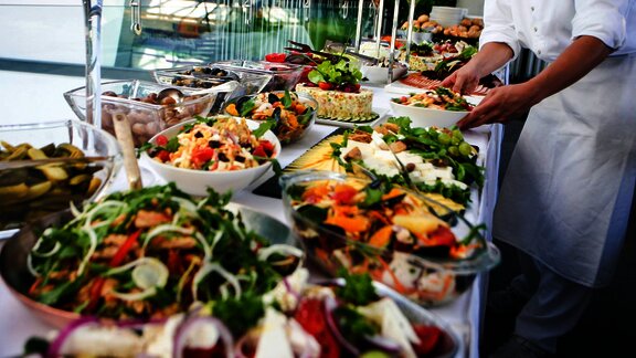 a buffet setup with a variety of fresh, colorful dishes, including salads, vegetables, and pickles. A person in a chef's uniform is arranging the food, ensuring everything is neatly displayed.