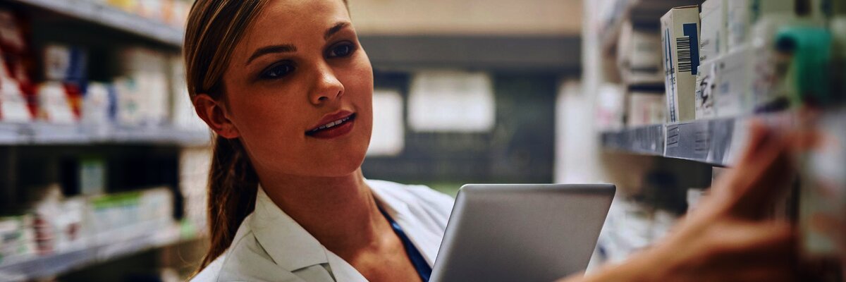 Female pharmacist organizing medications on a shelf while holding a tablet for inventory management in a pharmacy