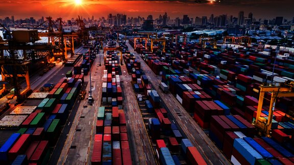 A busy container port with rows of colorful shipping containers, cranes, and cargo trucks, set against a city skyline at sunset, symbolizing global trade and logistics