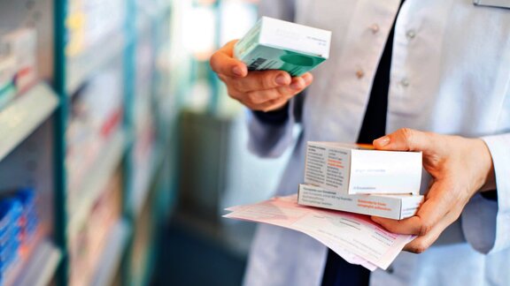 A pharmacist holds several boxes of medication and a prescription while organizing items on shelves in a pharmacy
