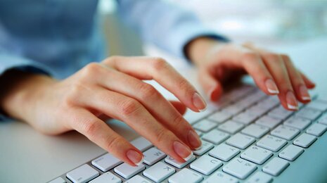 a woman's hands typing on a white computer keyboard, with a bright, softly blurred background